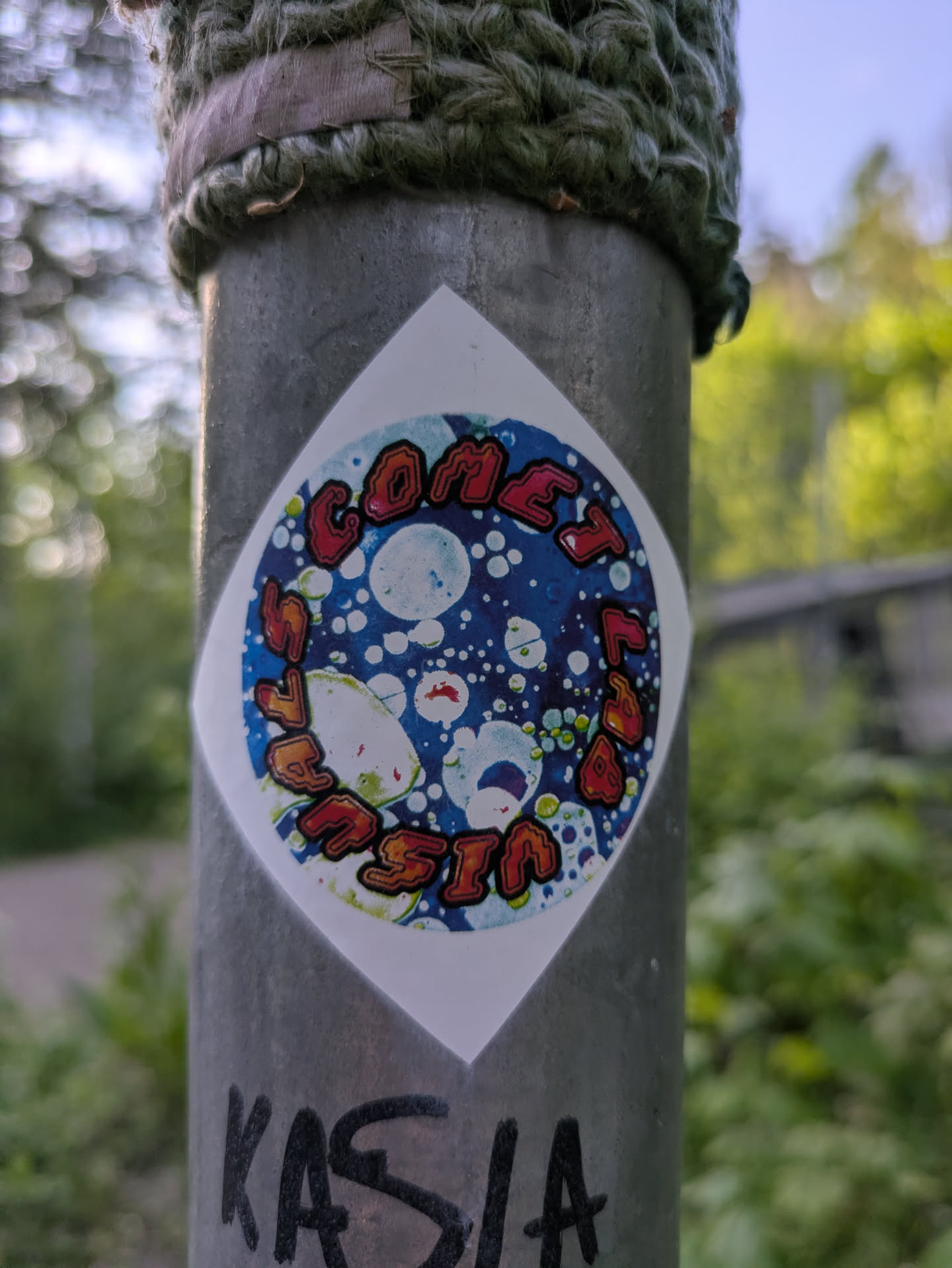This is a square sticker with rounded corners that features a circular design. Inside the circle, there's a celestial-themed illustration with a blue background, white bubble-like spheres, and a colorful outer ring with the text "Comet Tailed Invasion". The sticker is placed on a gray metal pole.