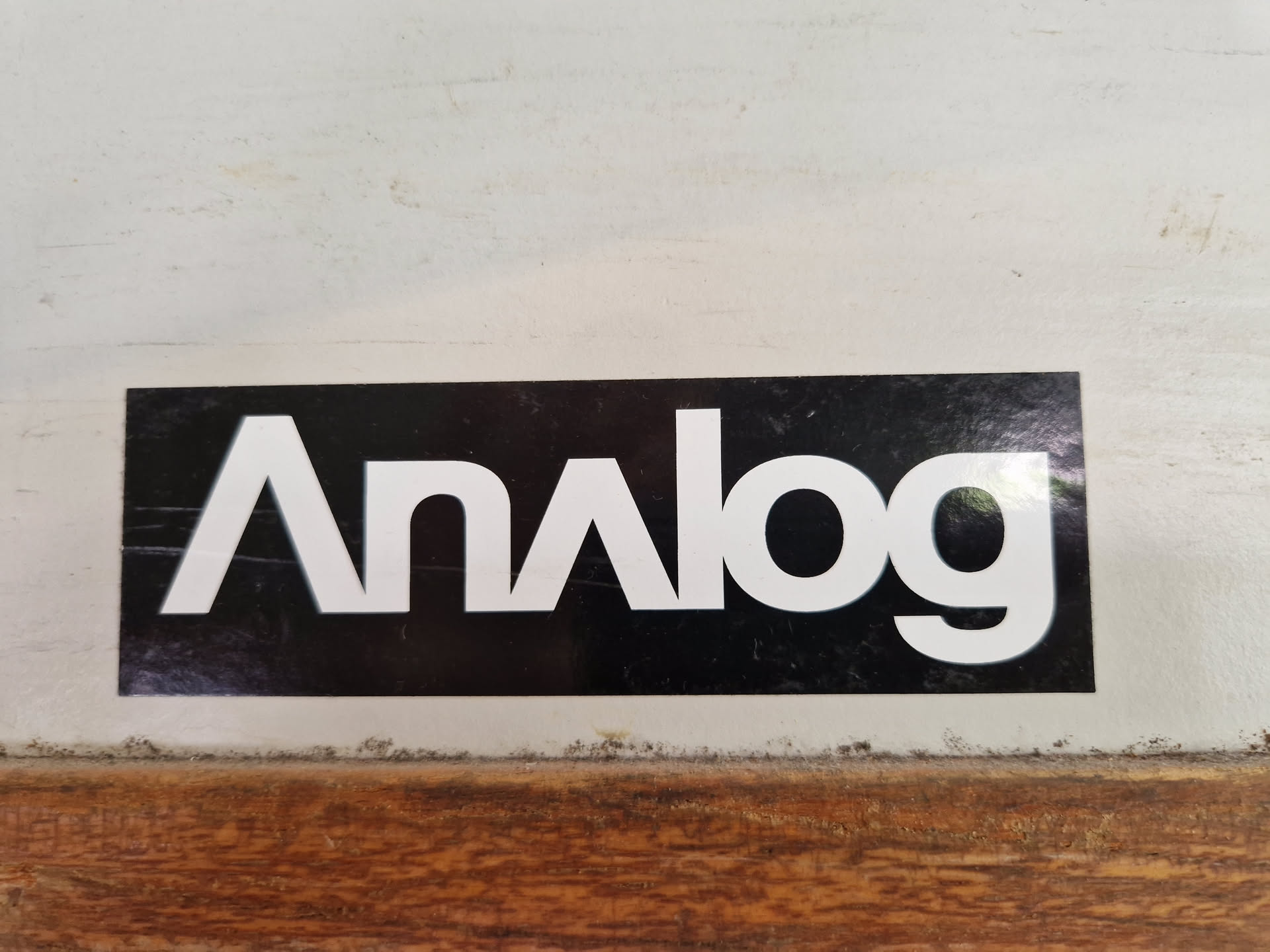 A black and white sticker with the word "Analog" written in a stylized font. The sticker is affixed to a light-colored surface that looks like painted wood or metal. The background is slightly dirty or worn.
