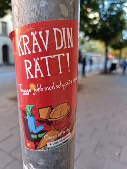 A red sticker advocating for fair wages and safe jobs. It features an illustration of two workers and text in Swedish.  The sticker is affixed to a metal pole.