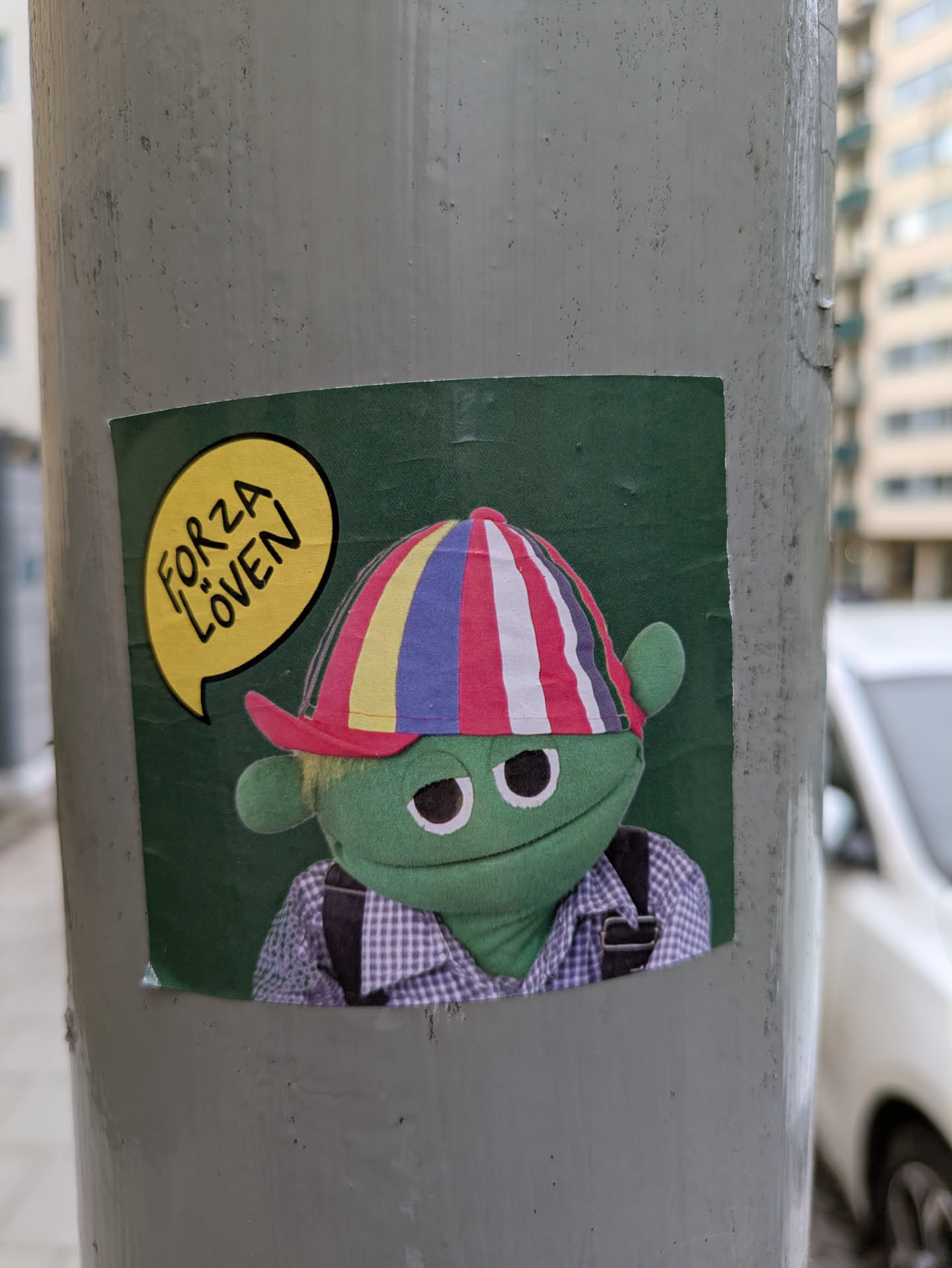 This sticker shows a green puppet with a slightly grumpy expression, wearing a cap with red, blue, yellow and white stripes. The puppet also has a light blue checked shirt with a black overall. Above the puppet is a yellow speech bubble with the text "FORZA LÖVEN". The background is green. The sticker is attached to a pole.