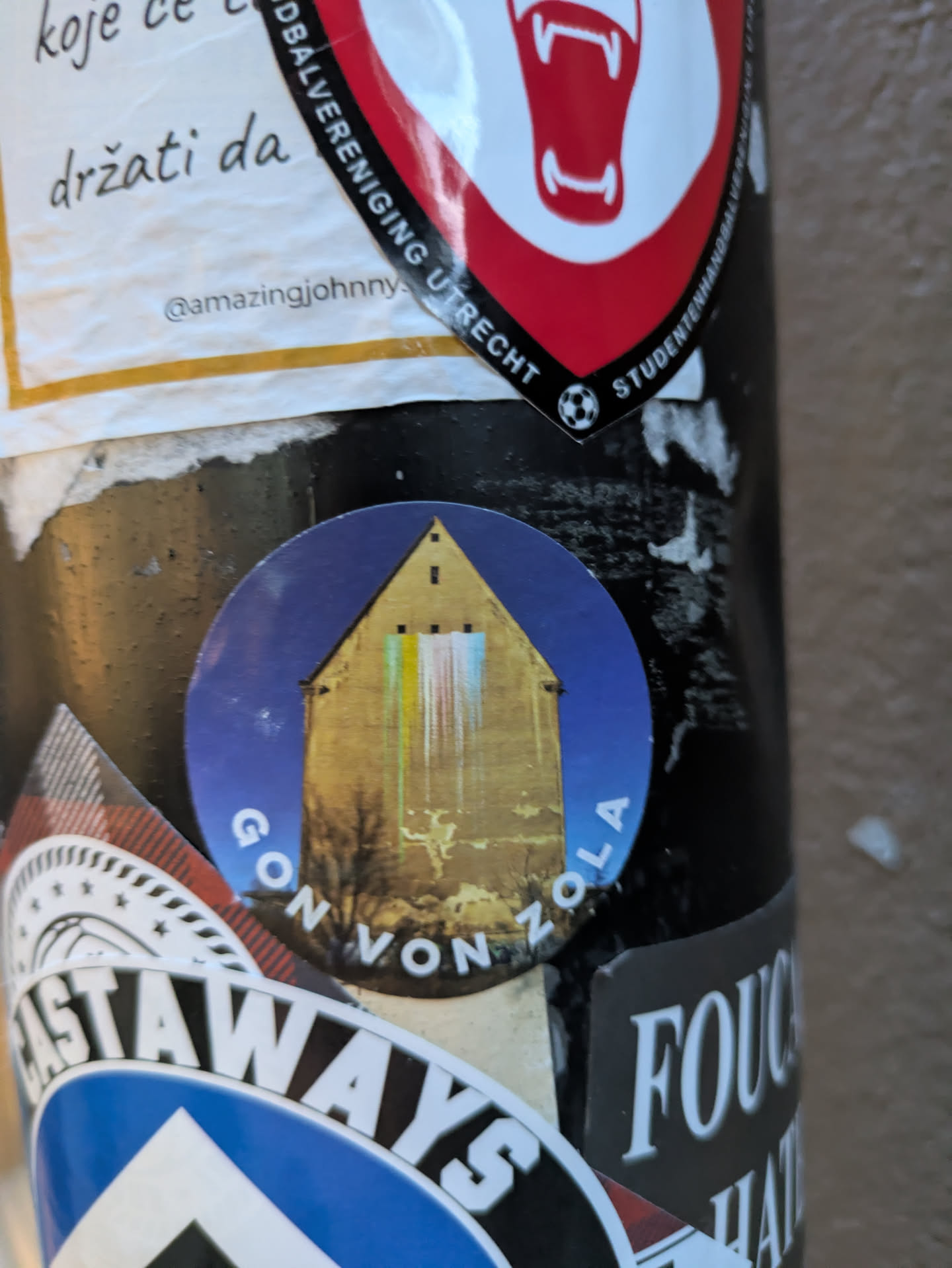 The sticker is circular with a deep blue background. It prominently displays a yellowish-tan building with a steep, peaked roof and several visible windows. A colorful, abstract vertical stripe pattern, resembling light or rain, is projected onto the building's facade. The text 'GON VON ZOLA' is written in white capital letters in a curved arc across the bottom of the sticker. The sticker appears to be slightly worn, adding to its character.