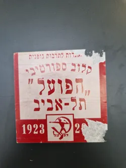A weathered, red and white sticker with Hebrew text and a symbol reminiscent of socialist or communist imagery.  The numbers 1923 and 20 are visible, suggesting a historical context. The sticker shows signs of age and wear.