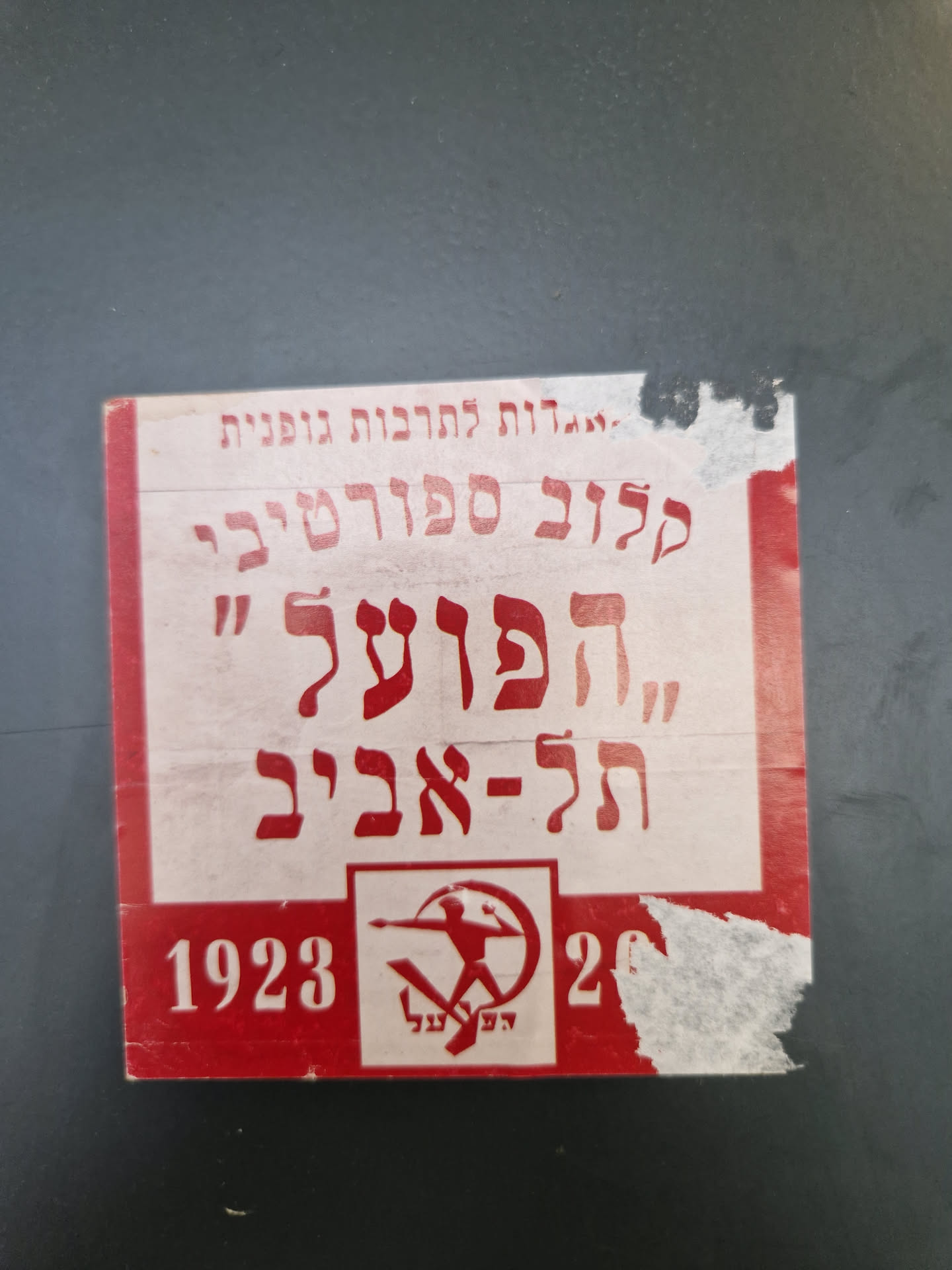 A weathered, red and white sticker with Hebrew text and a symbol reminiscent of socialist or communist imagery.  The numbers 1923 and 20 are visible, suggesting a historical context. The sticker shows signs of age and wear.