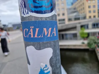 A blue sticker with the word CÄLMÄ (Swedish for Calm) in pink lettering is seen on a pole in an urban setting.  The background is blurry, showcasing a city street and canal.