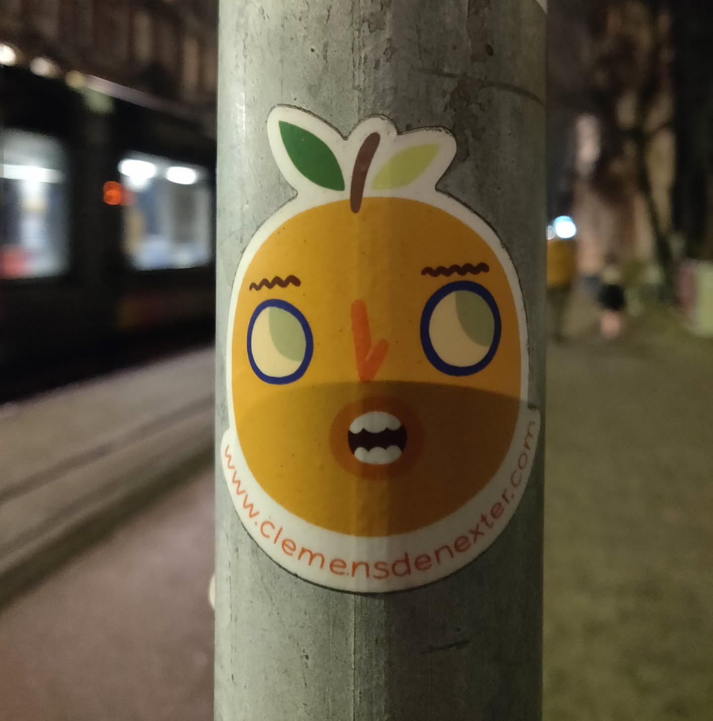 This sticker features a cartoon-like illustration of an orange with a surprised expression. It's a vibrant yellow-orange with blue-rimmed eyes and a simple, friendly design.  The sticker is affixed to a metal pole at night, with a blurred tram visible in the background.  The website address www.clemensdenexter.com is displayed at the bottom of the design.