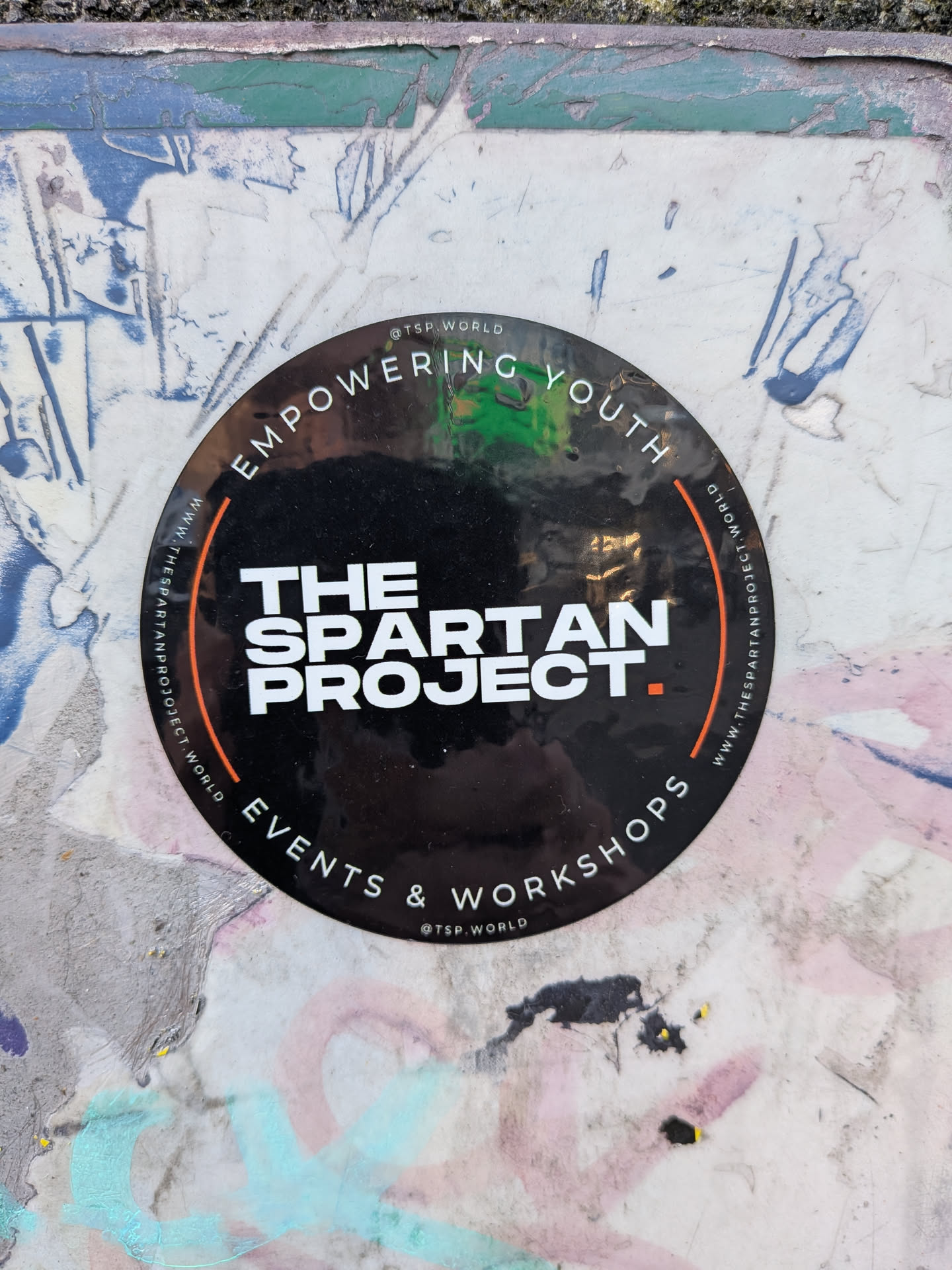 A circular black sticker featuring bold white text. The center prominently displays 'THE SPARTAN PROJECT.' with a small orange square acting as a period. Along the top curved edge, 'EMPOWERING YOUTH' is written in white capital letters, and along the bottom curved edge, 'EVENTS & WORKSHOPS' is displayed. The website 'WWW.THESPARTANPROJECT.WORLD' is printed twice, following the inner curve on both the left and right sides. The social media handle '@TSP.WORLD' appears at the very top and bottom of the sticker, outside the main circular text arcs. The sticker has a slightly glossy finish, showing some reflections and minor wear, indicating it's adhered to a textured, possibly painted, surface.