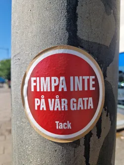A red round sticker on a grey pole with the text "FIMPA INTE PÅ VÅR GATA" which translates to "Don't butt out on our street" in Swedish. At the bottom it says "Tack" which means "Thanks".