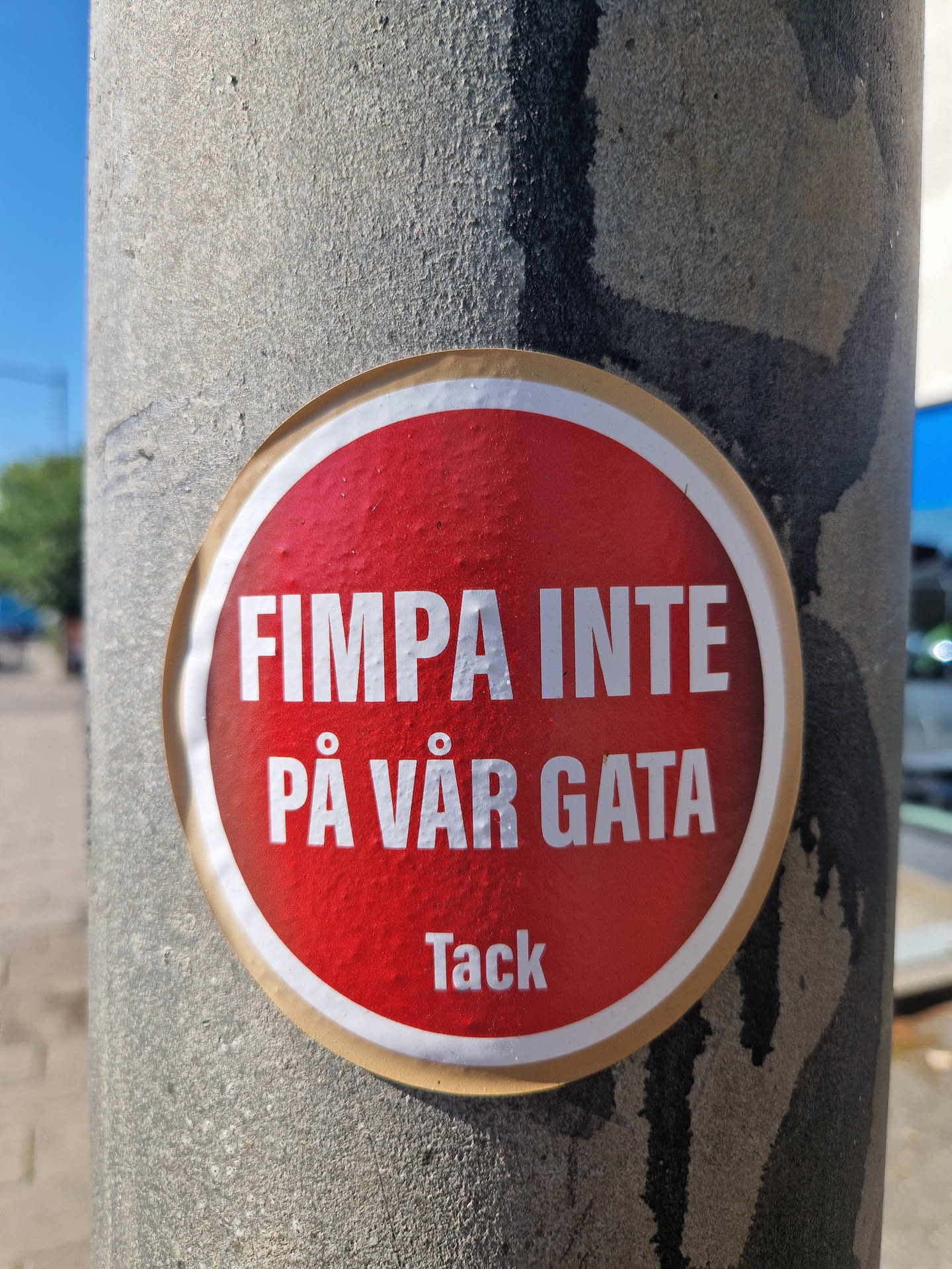 A red round sticker on a grey pole with the text "FIMPA INTE PÅ VÅR GATA" which translates to "Don't butt out on our street" in Swedish. At the bottom it says "Tack" which means "Thanks".
