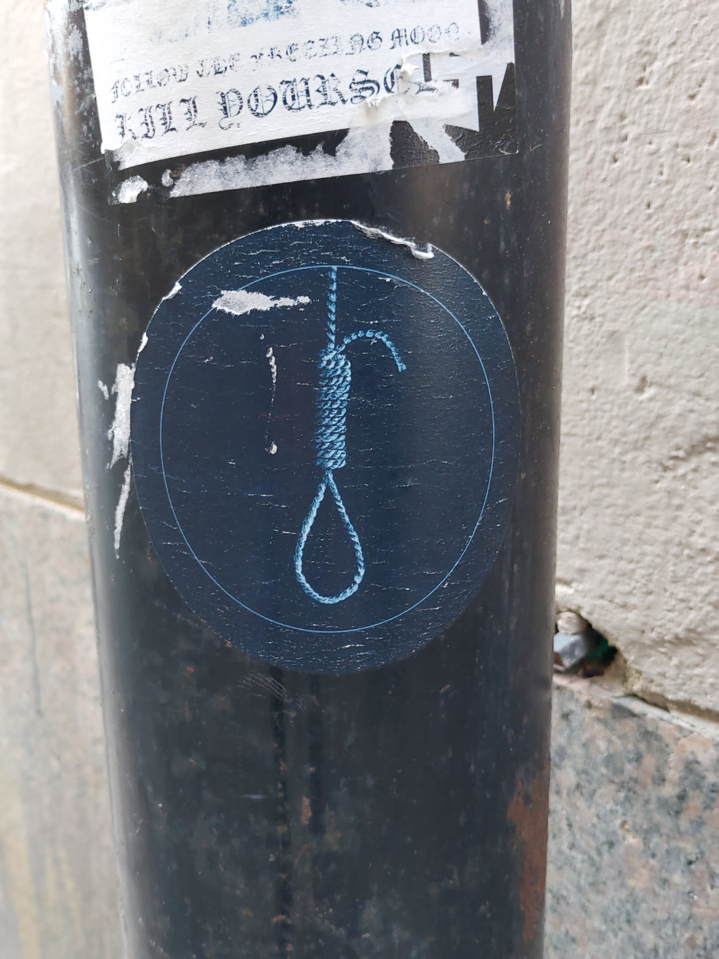 A round dark blue sticker with a light blue outline depicts a noose. The sticker is affixed to a dark metal pole. Another sticker above it is partially visible, with some illegible text. The background is a light beige wall.