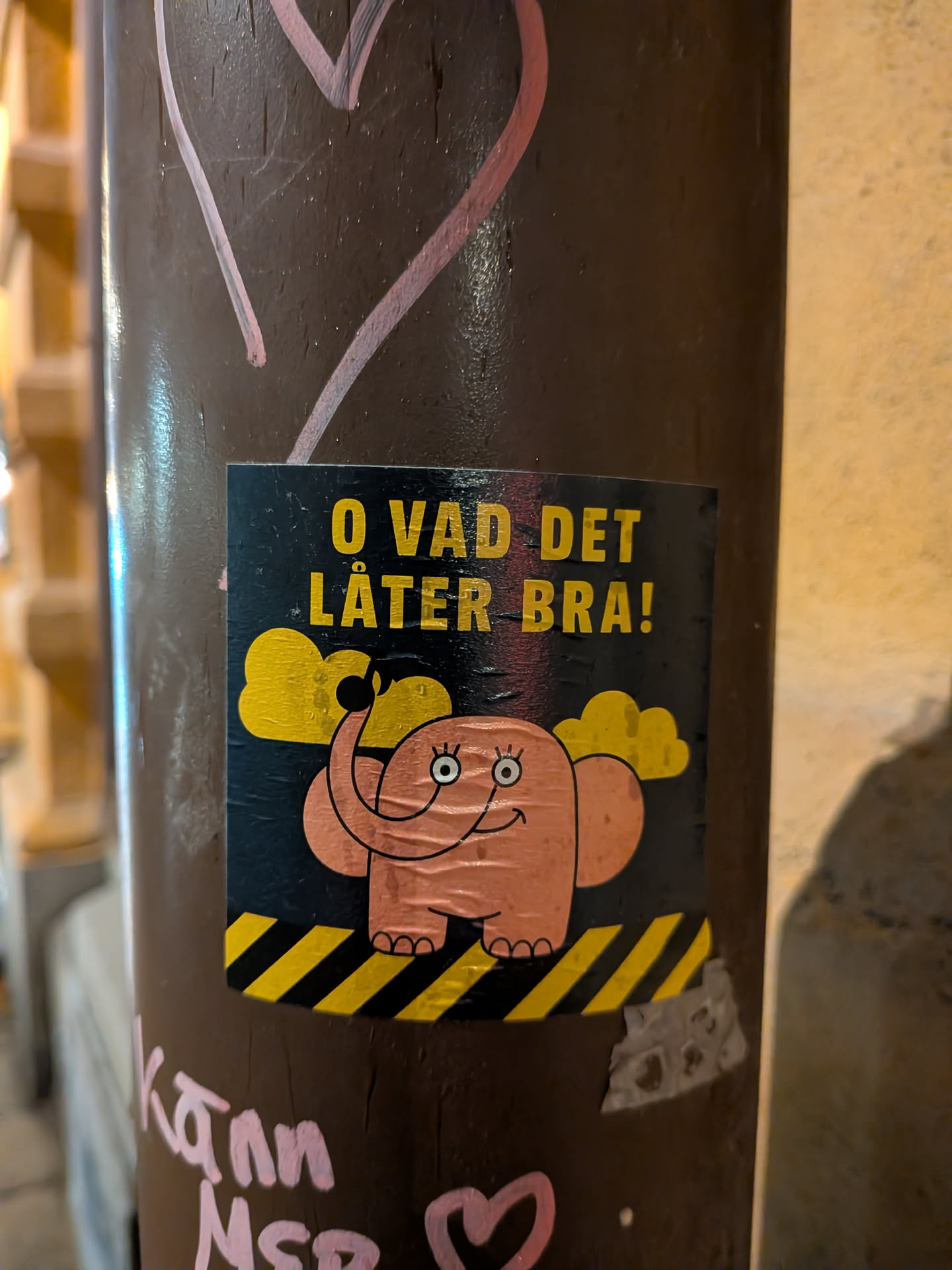 The sticker is square-shaped with a dark blue/black background. In the center is a cartoonish pink elephant with large, round eyes, a wide smile, and rosy cheeks. It has its trunk raised and appears to be waving. Above the elephant are two fluffy yellow clouds. The bottom of the sticker features a pattern of diagonal yellow and black stripes, reminiscent of hazard tape. The text 'O VAD DET LÅTER BRA!' is prominently displayed in yellow, blocky letters at the top of the sticker.