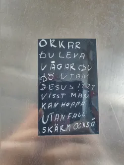 A black sticker with white handwritten text in Swedish. The text seems to be a philosophical or religious statement questioning life, death, and faith. It poses questions about the strength to live and the courage to die without religious belief, suggesting that one might take risks even without safety.