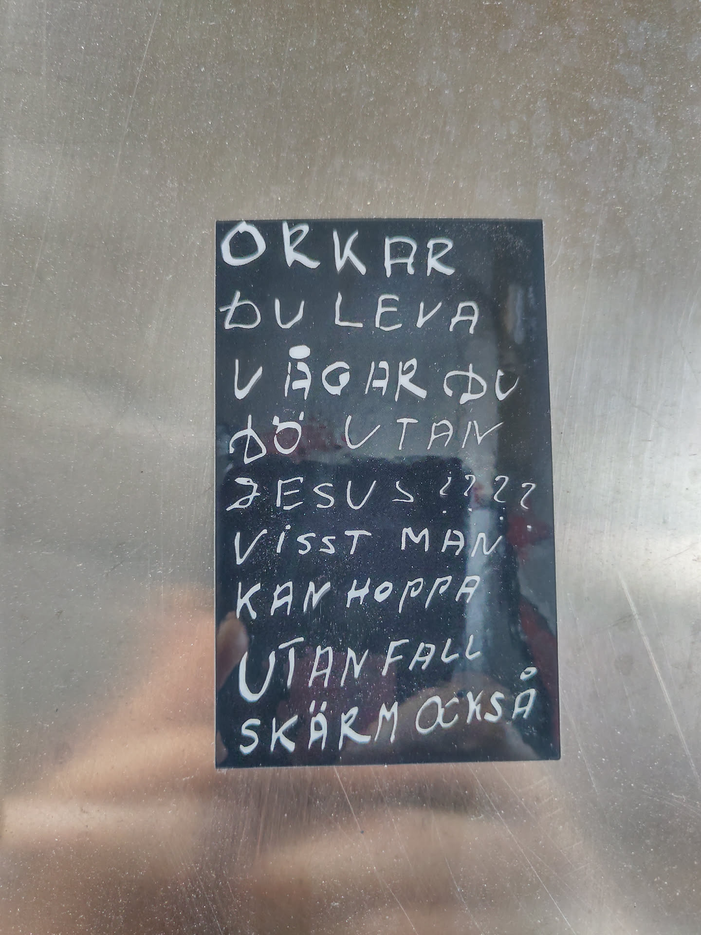 A black sticker with white handwritten text in Swedish. The text seems to be a philosophical or religious statement questioning life, death, and faith. It poses questions about the strength to live and the courage to die without religious belief, suggesting that one might take risks even without safety.