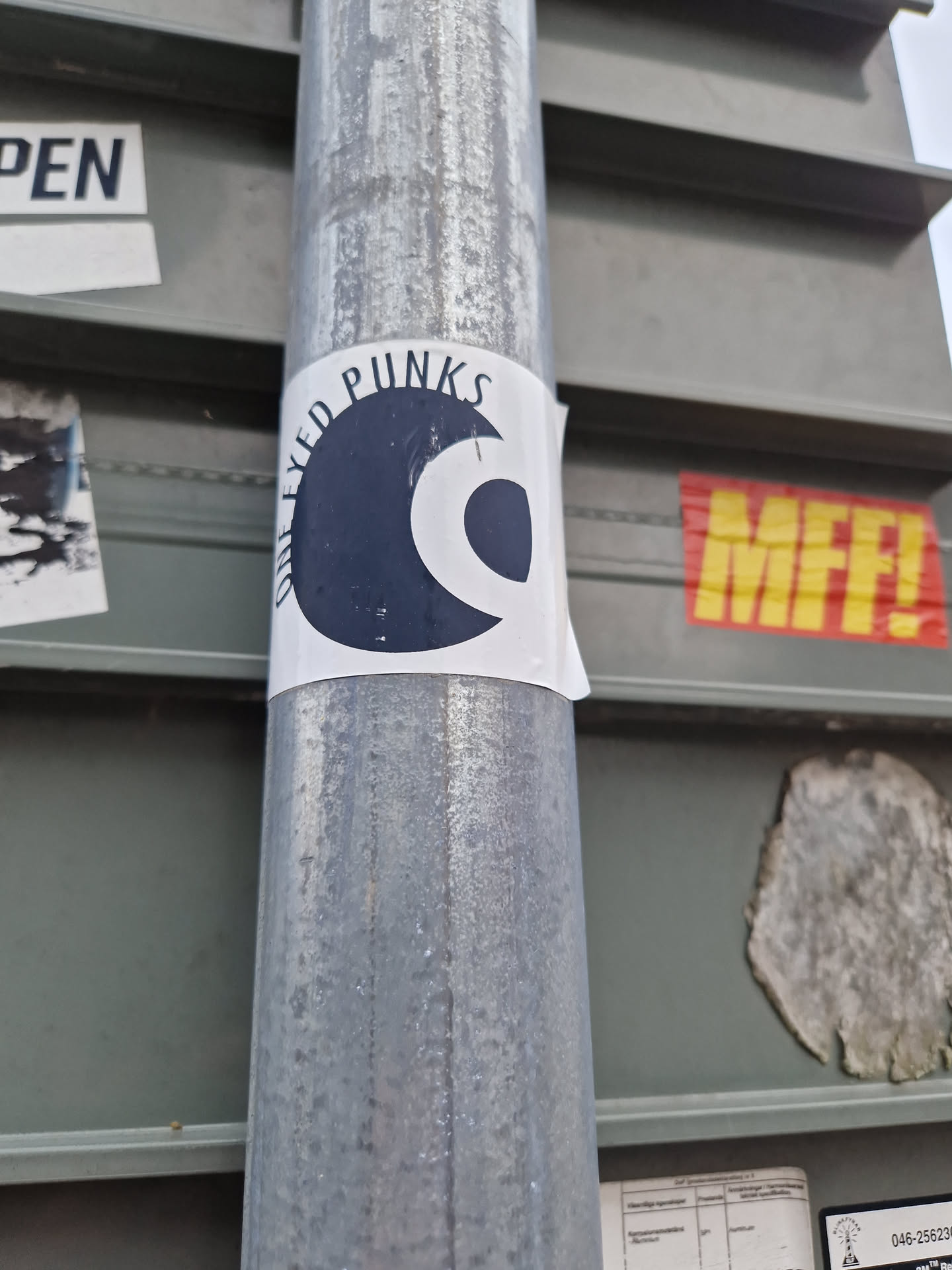 A round sticker with a dark blue/black design featuring a stylized eye and the text "One Eyed Punx" printed in a circular pattern around the main graphic. The sticker is affixed to a metal pole.
