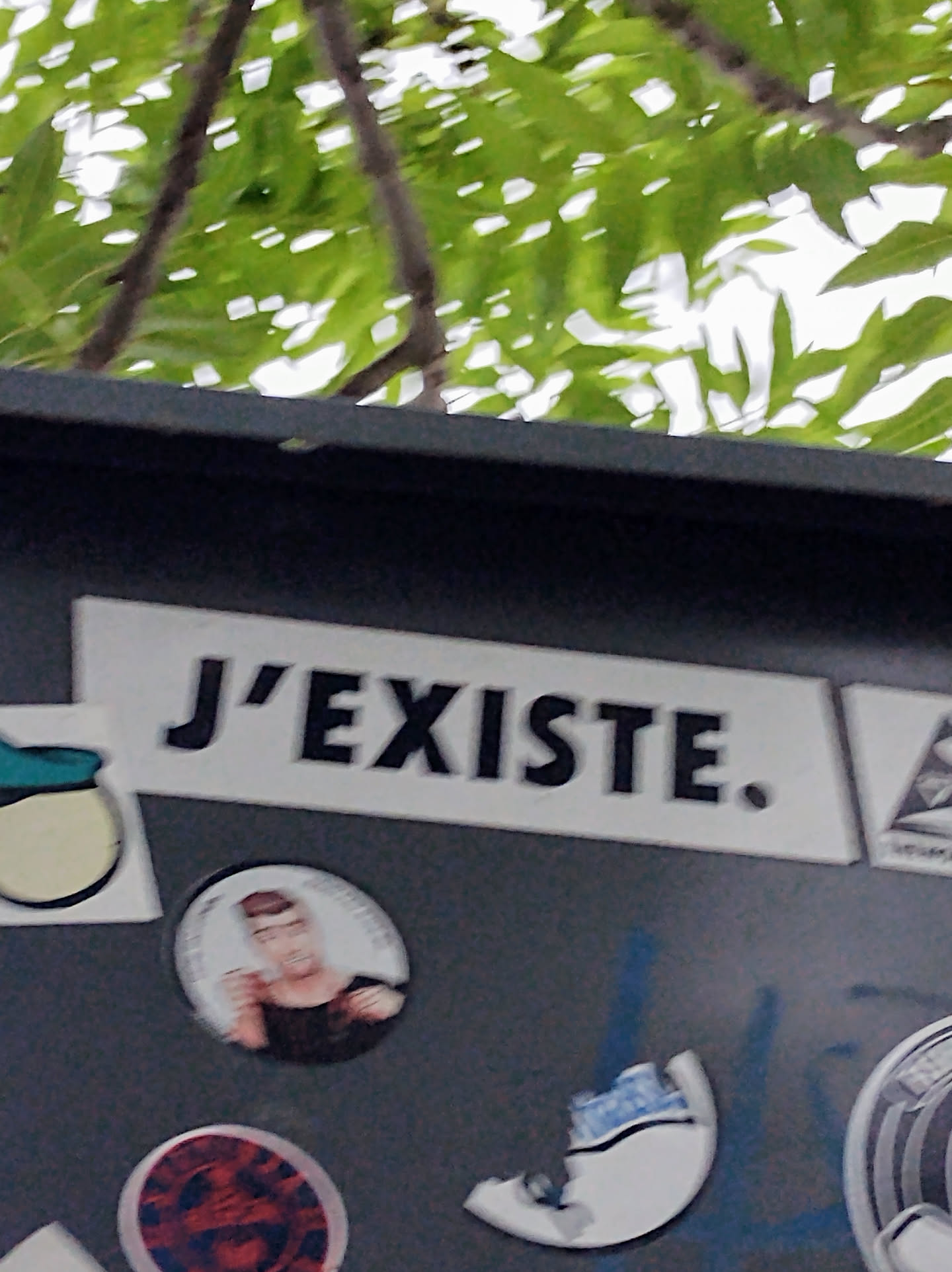 A black and white sticker with the French phrase "J'existe." (I exist) on a gray surface. The sticker is surrounded by other stickers and graffiti.