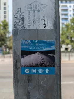The sticker features an image of sand dunes under a blue sky, with a person standing in the distance. The word "GEOGRAFIA" is printed at the top of the image and a Spotify code at the bottom.
