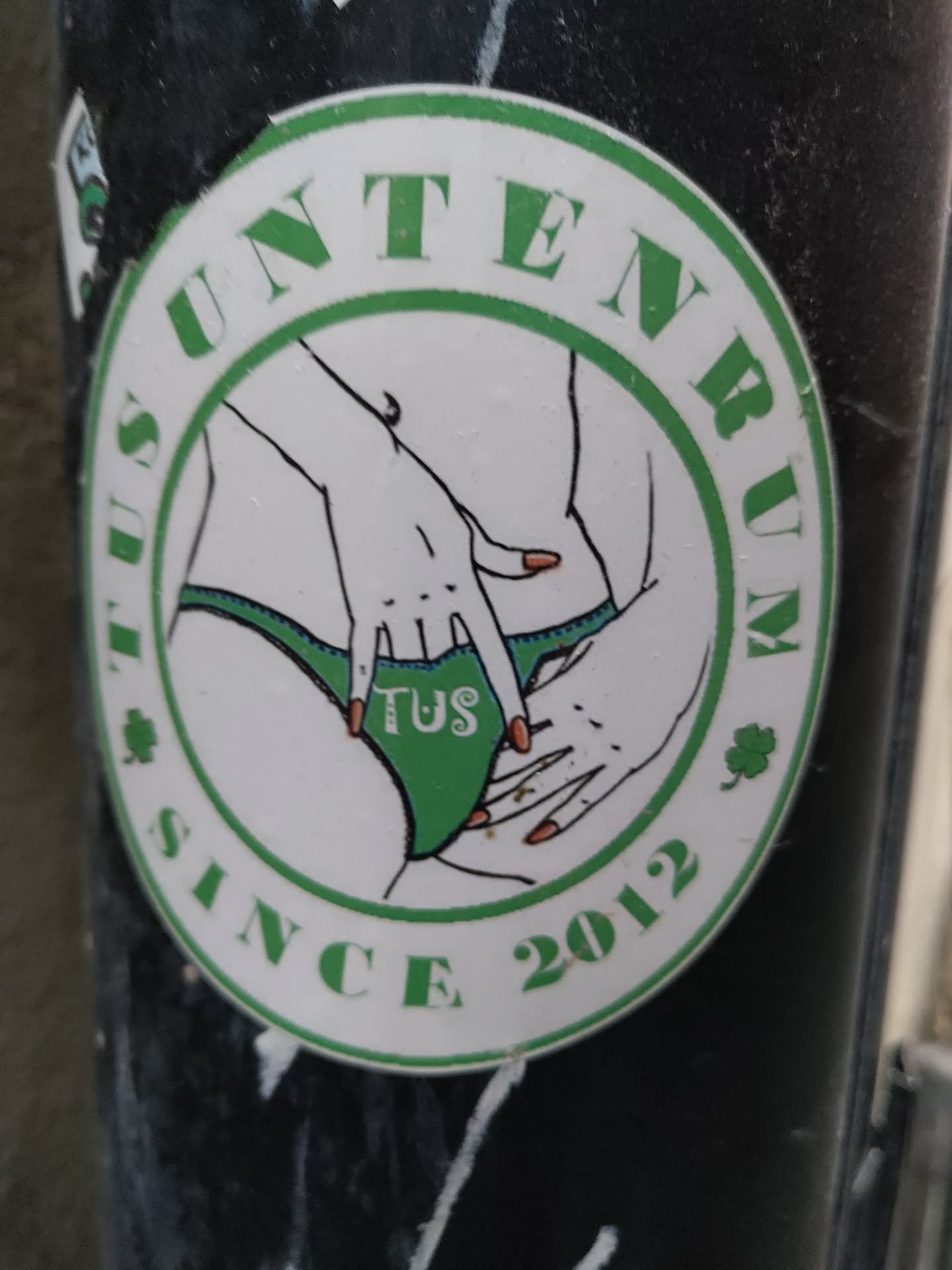 A round sticker with a green border featuring an illustration of a woman's lower body and hands. The text "Tus Unternum since 2012" is written around the image.
