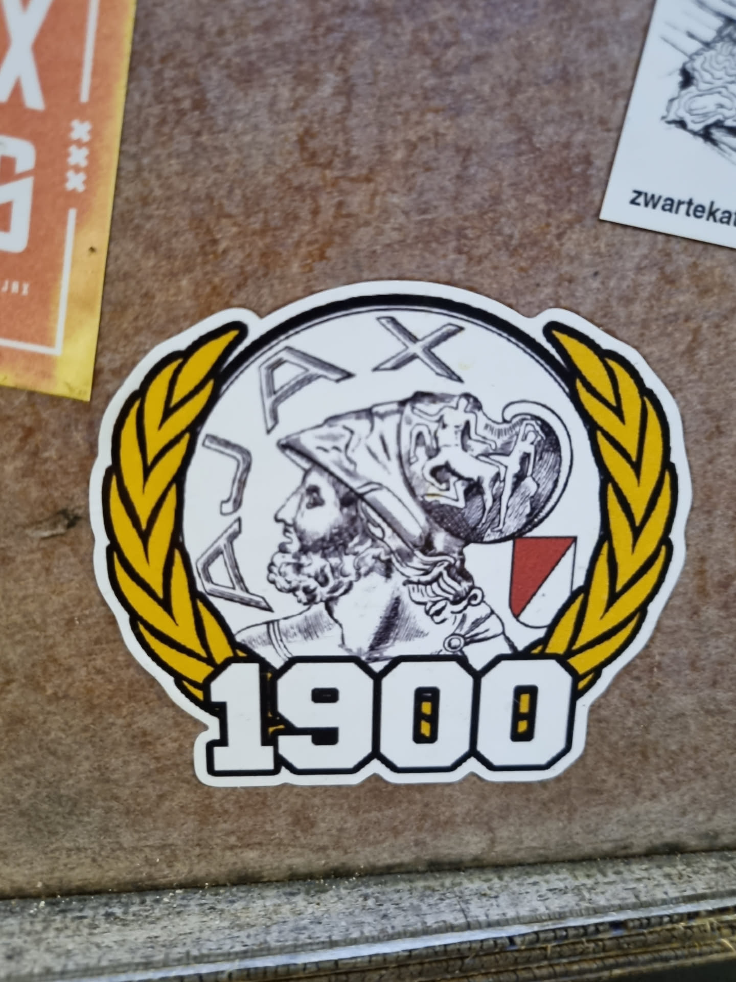 A sticker featuring the Ajax Amsterdam logo, which includes a laurel wreath, the year 1900, and a depiction of a warrior's head. The style is vintage and classic.