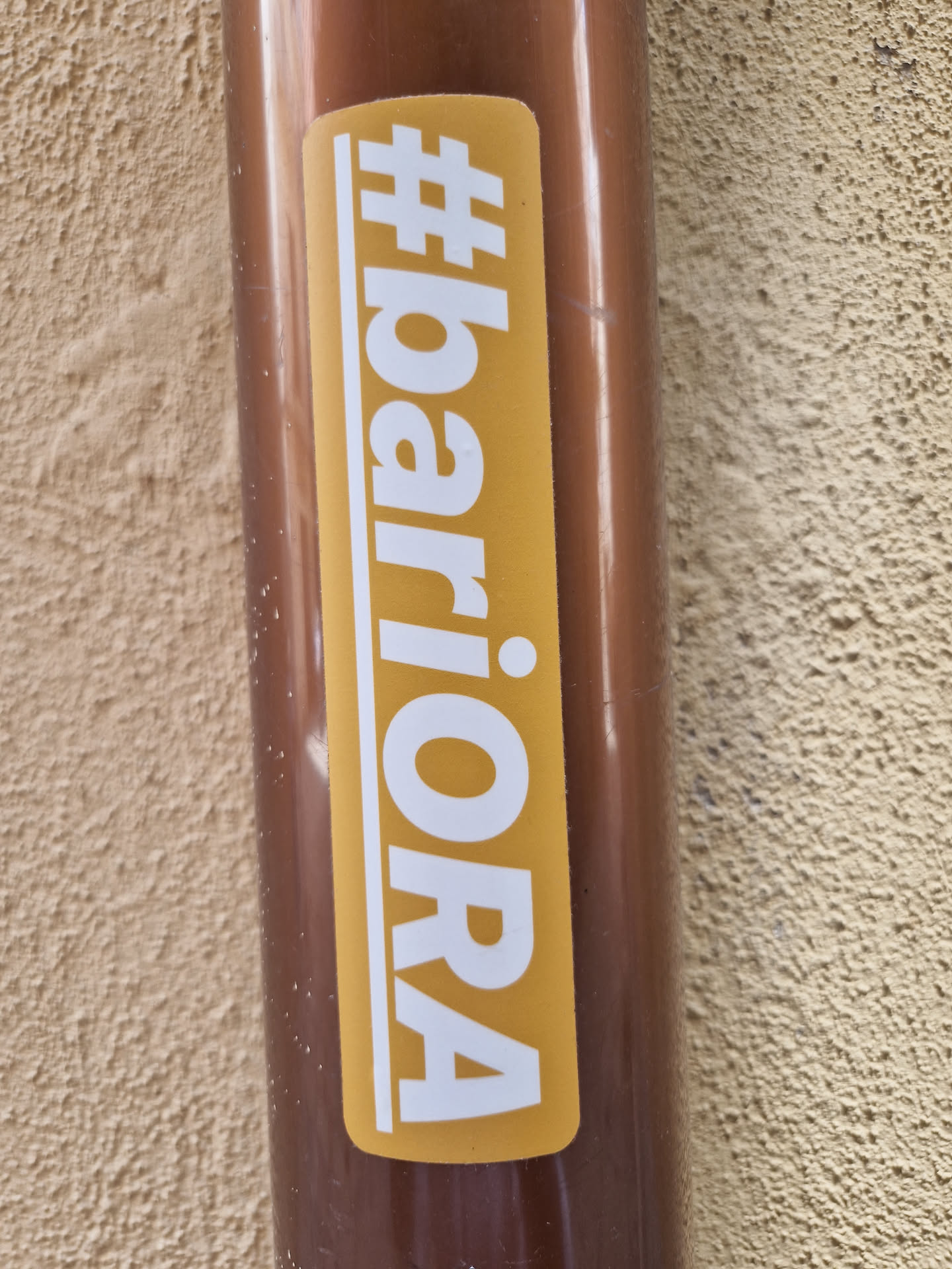 A mustard yellow-colored sticker with the text '#bariora' in white, uppercase letters.  The hashtag is included as part of the design. The sticker is affixed to a brown cylindrical object, possibly a pipe.