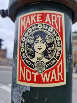 A sticker featuring a woman's face surrounded by roses and the text 'Make Art Not War'. The sticker is attached to a dark green pole.