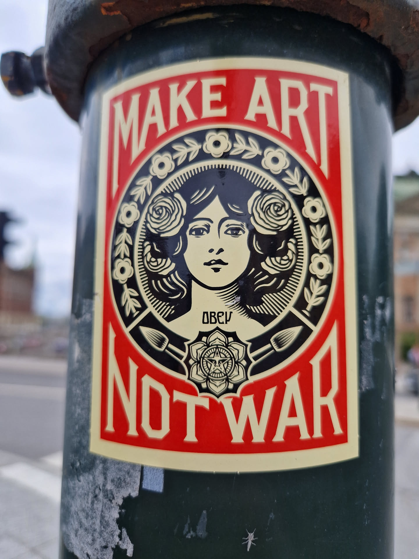 A sticker featuring a woman's face surrounded by roses and the text 'Make Art Not War'. The sticker is attached to a dark green pole.
