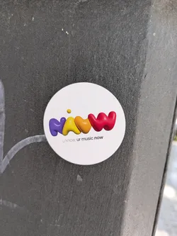 Street sticker A round, white sticker adhered to a textured, dark surface features the brand name 'NAWW' in a stylized, colorful design. The letters are rendered in rounded, three-dimensional shapes with a glossy finish, transitioning from purple to yellow to orange and finally to red. A small yellow dot floats above the first 'W', accentuating the playful feel of the logo. Beneath the brand name, the tagline 'u know, ur music now' is printed in a smaller, sans-serif font. The sticker's clean design and vibrant colors contrast with the rough, dark background.