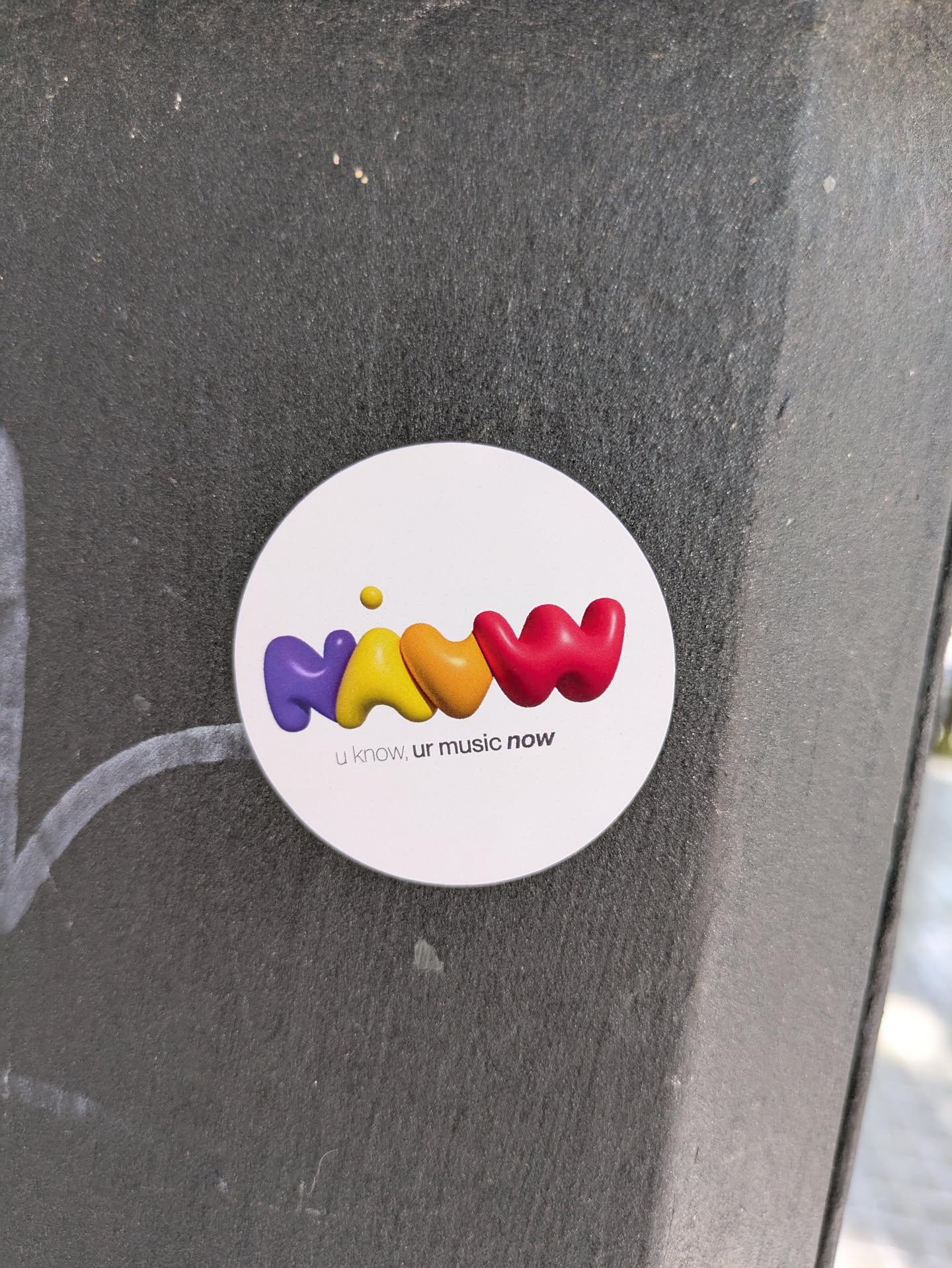 A round, white sticker adhered to a textured, dark surface features the brand name 'NAWW' in a stylized, colorful design. The letters are rendered in rounded, three-dimensional shapes with a glossy finish, transitioning from purple to yellow to orange and finally to red. A small yellow dot floats above the first 'W', accentuating the playful feel of the logo. Beneath the brand name, the tagline 'u know, ur music now' is printed in a smaller, sans-serif font. The sticker's clean design and vibrant colors contrast with the rough, dark background.