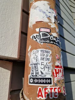 A sticker featuring a hand-drawn illustration of a person, text that reads "The Miserable Youth of Our Time" and "5/4! (2024)", and a Spotify code. The style is reminiscent of indie music or zine culture. 