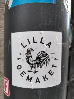 A white sticker with a black rooster illustration and text. The text says "LILLA GEMÄKET" around the rooster. The rooster is stylized and detailed.