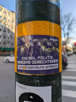 A sticker on a pole with a picture of a group of people protesting, text in Dutch saying "Everywhere police, nowhere justice. Tolerate no police intimidation", and a website address at the top right corner.  The sticker has a yellow border.