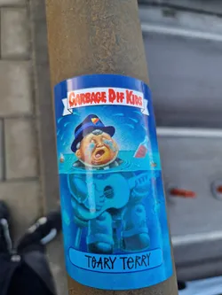 A vintage Garbage Pail Kids sticker featuring Teary Terry, a character submerged in water while playing the guitar. The sticker has a blue background with some water splash illustrations. The name of the character is written at the bottom. The words Garbage Pail Kids are written on the banner at the top of the sticker.