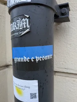 A rectangular sticker featuring a prominent horizontal blue band across its upper half, set against a black background. White text in a distinctive gothic or blackletter font reads 'grande e pesante'. The sticker shows some wear, minor bubbling, and is adhered to a textured, wet surface.