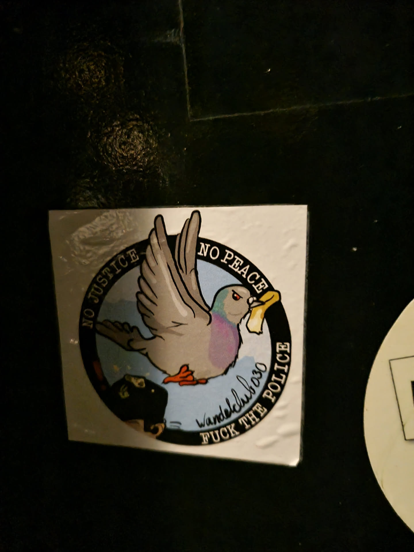 A sticker featuring a cartoon pigeon carrying a piece of paper in its beak. The text around the pigeon says "No Justice, No Peace" and "Fuck the Police." There's also a signature that reads "Wandelclub030."