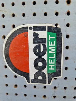 A weathered sticker from the brand Poeri, advertising their helmets. The sticker features a red and black design with the brand name prominently displayed.  The word "HELMET" is visible in green text. The sticker shows its age with some dirt and wear. It is attached to a gray pegboard.