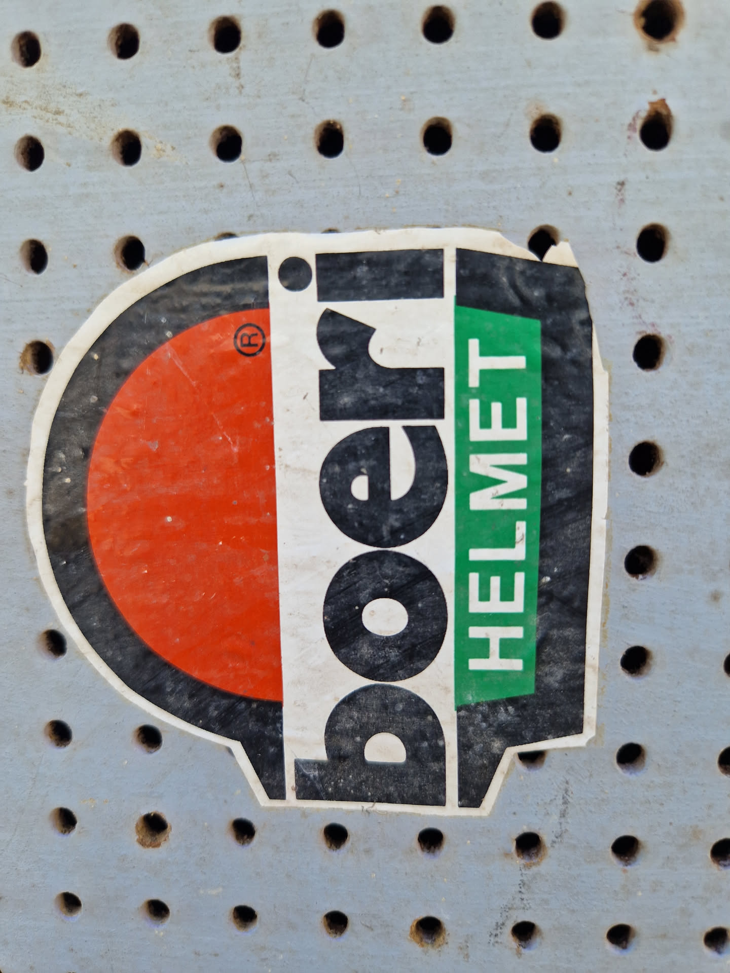 A weathered sticker from the brand Poeri, advertising their helmets. The sticker features a red and black design with the brand name prominently displayed.  The word "HELMET" is visible in green text. The sticker shows its age with some dirt and wear. It is attached to a gray pegboard.