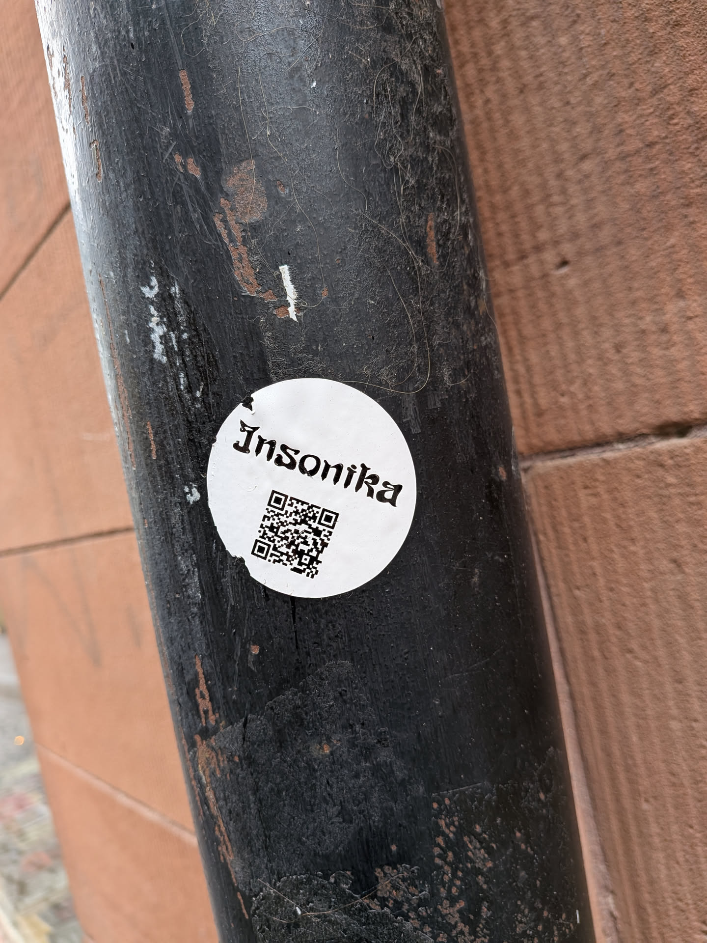 A white, circular sticker affixed to a dark, weathered pole. The top half of the sticker features the word "Insonika" in a distinctive blackletter (gothic) font, rendered in black. Below the text, a black QR code is centrally placed. The sticker shows minor wear and tear, particularly around its edges, suggesting it's a physical sticker exposed to the elements.