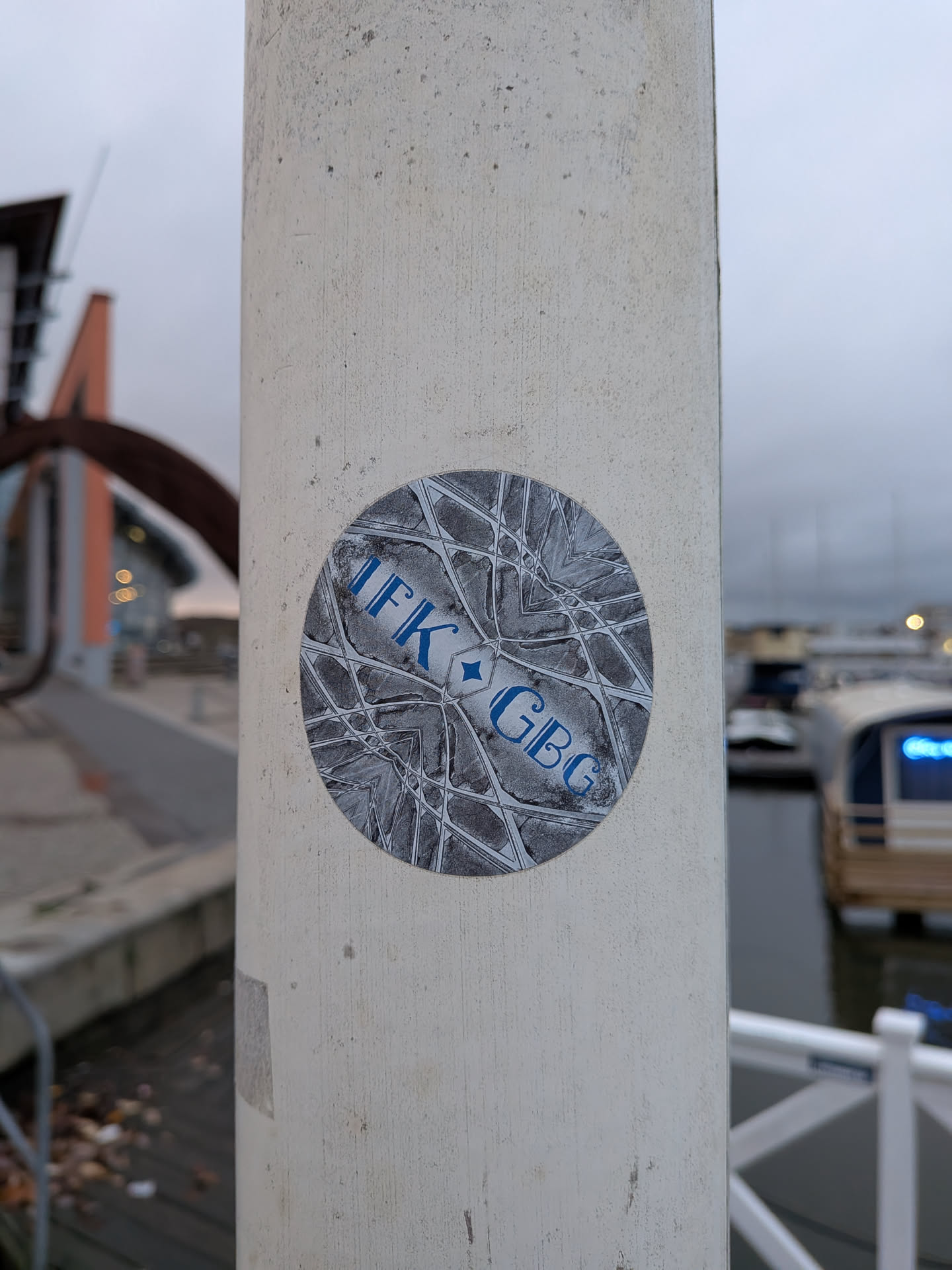 The sticker is circular and features a complex, symmetrical geometric pattern in shades of gray and white. Superimposed on this pattern, in bold blue lettering, are the letters 'IFK' on the left and 'GBG' on the right, separated by a small blue diamond shape. The overall design is abstract and stylized, with the text clearly legible against the intricate background.