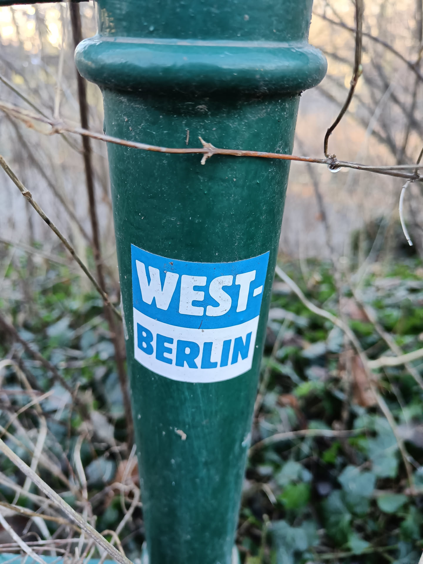 A close-up shot of a sticker on a dark-green pole. The sticker is rectangular with a blue top section reading "WEST-" in white uppercase lettering and a white bottom section reading "BERLIN" in uppercase lettering. The pole appears to be outdoors and surrounded by vegetation.