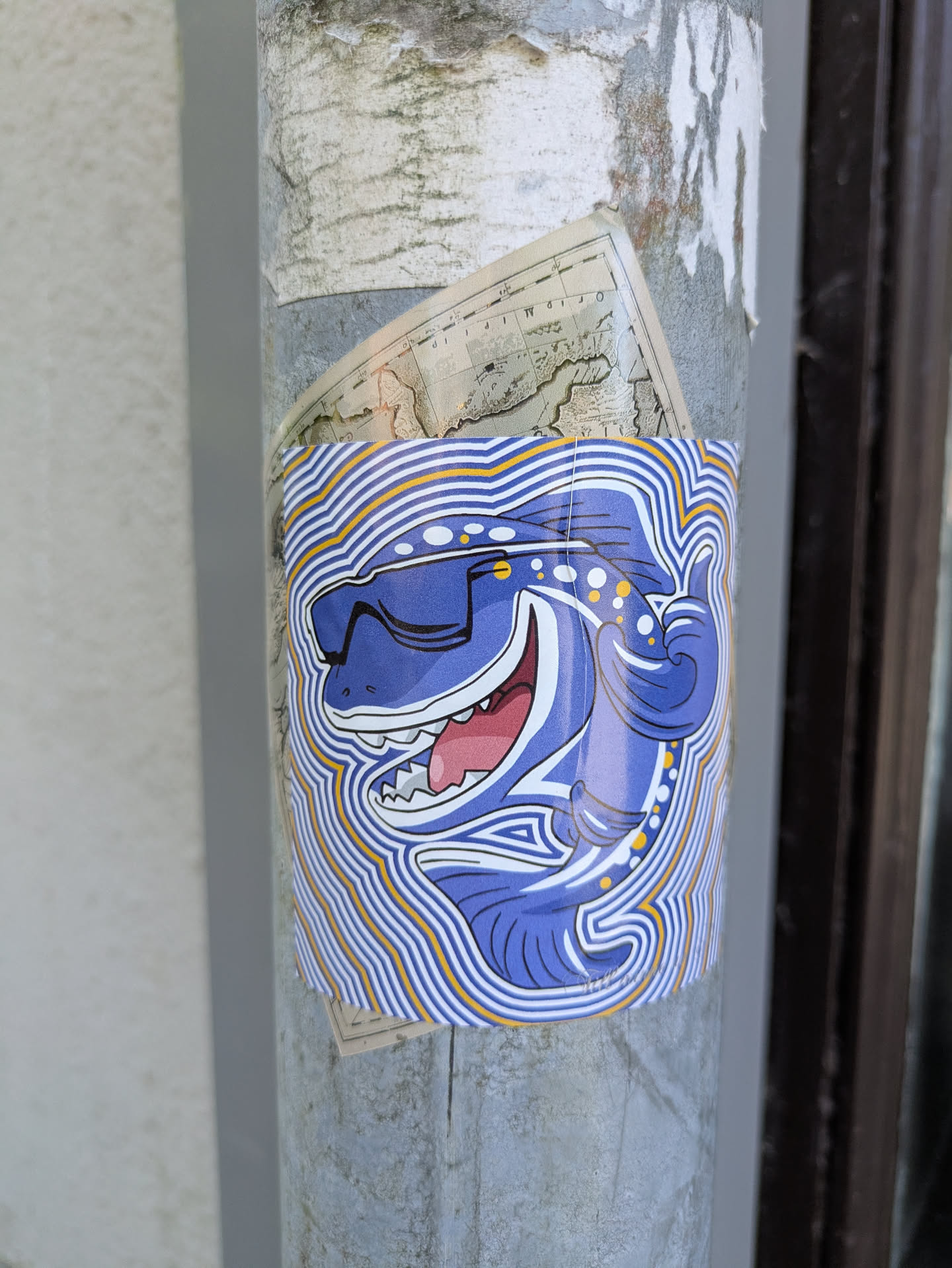 A sticker of a cool shark with sunglasses is affixed to a pole. The shark is blue with white spots and a big smile, set against a background of wavy blue and yellow lines. The style is cartoonish and playful. Another older sticker is behind it.