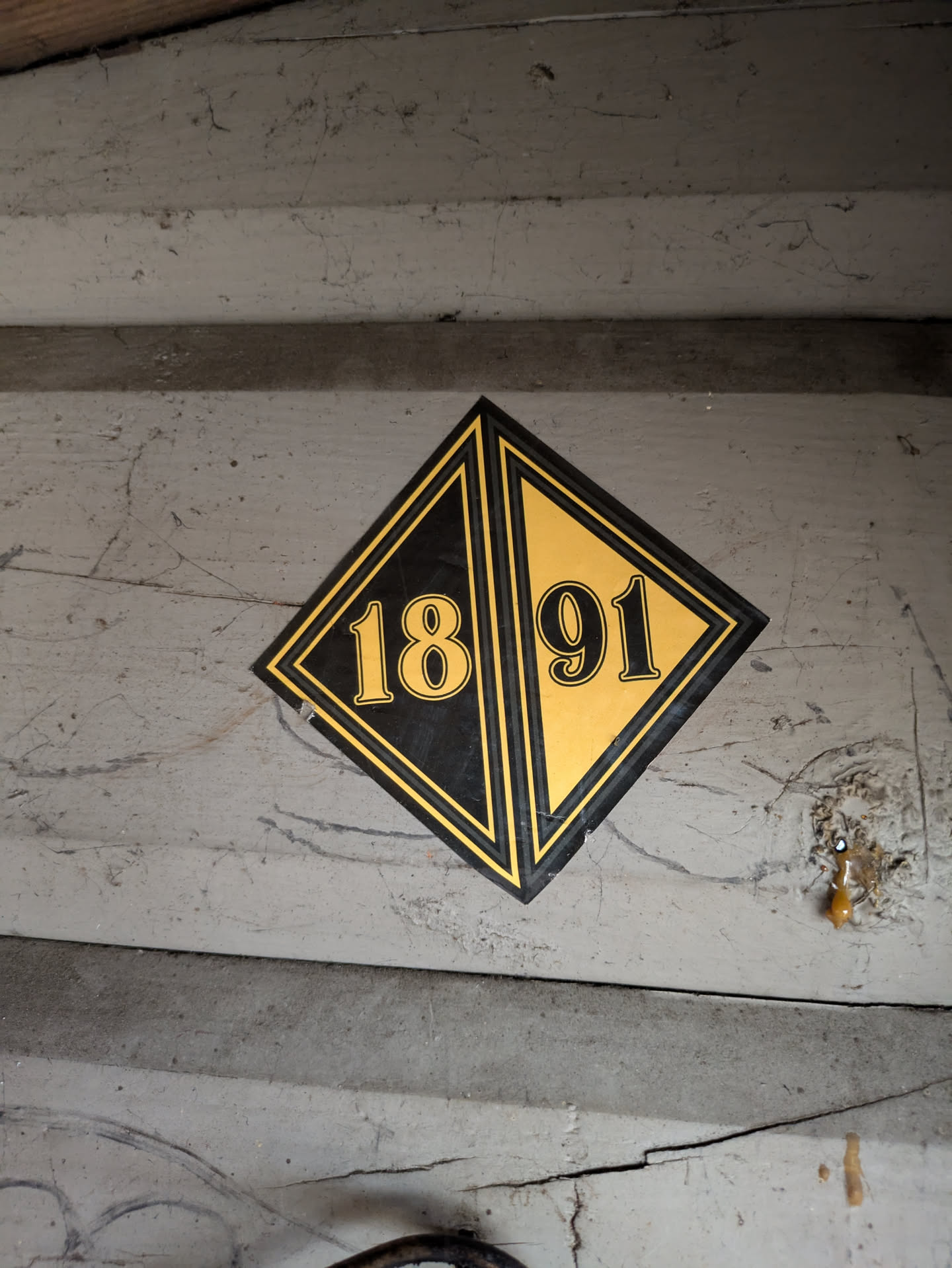 The image features a diamond-shaped sticker centrally placed on a light grey, worn, possibly wooden surface. The sticker is divided diagonally, with the top-left half being black and the bottom-right half being a golden yellow. The number '18' is prominently displayed in golden yellow on the black half, and '91' is in black on the golden yellow half, completing the year '1891'. The sticker has a double border, an inner black line and an outer golden yellow line. The background surface shows signs of age, including cracks, dirt, and scuff marks, suggesting a historical or aged context for the sticker.