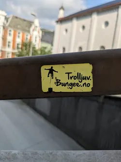 A rectangular yellow sticker with slightly peeling edges is affixed to a dark, textured metal railing. The sticker displays a black silhouette of a person in mid-air, suspended by a line, evoking a bungee jump. To the right of the silhouette, the words "Trolljuv." and "Bungee.no" are written in a casual, handwritten-like black font. The background of the image is blurred, showing architectural elements of buildings under a partly cloudy sky, suggesting an urban or scenic outdoor location.