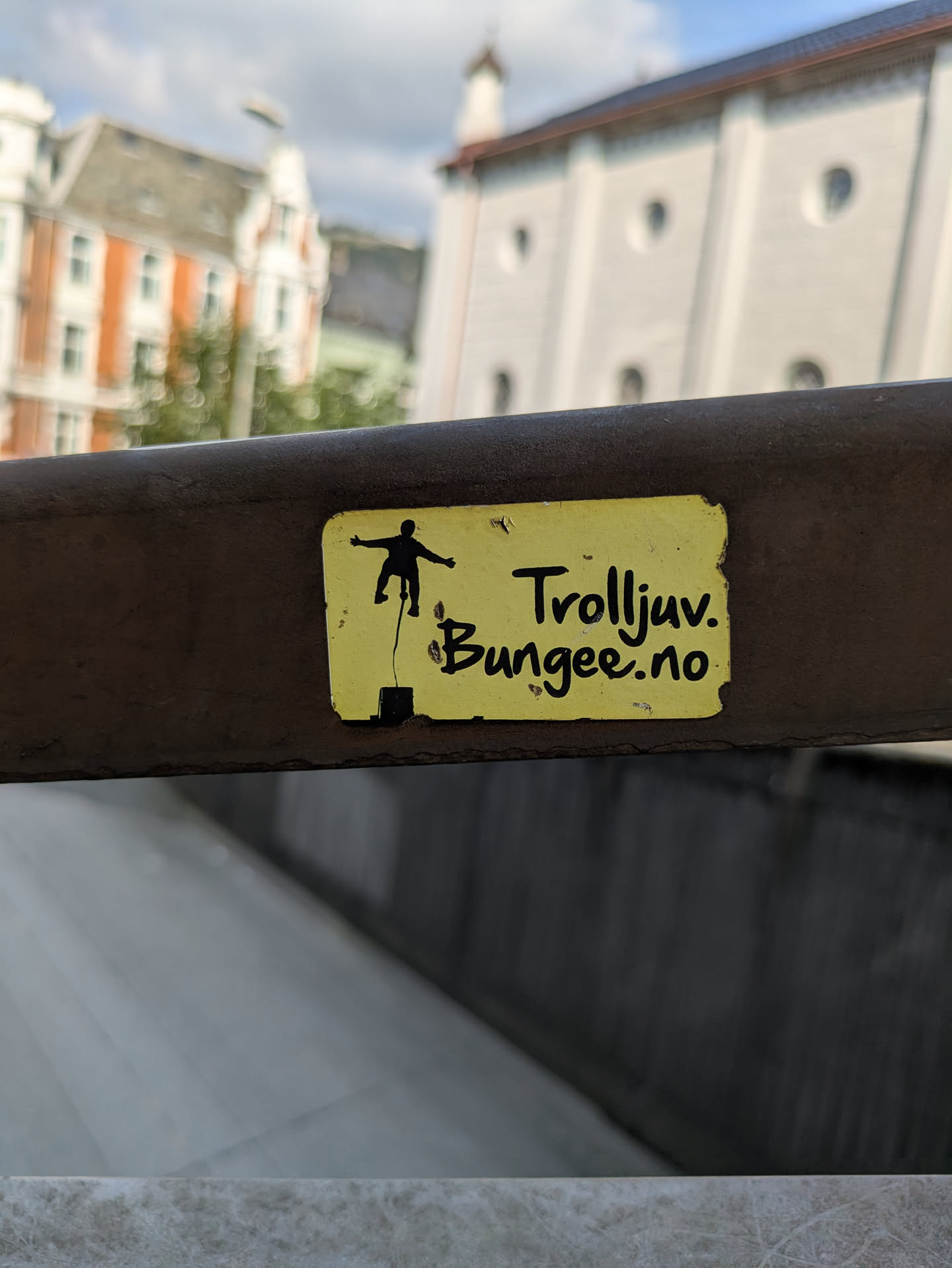 A rectangular yellow sticker with slightly peeling edges is affixed to a dark, textured metal railing. The sticker displays a black silhouette of a person in mid-air, suspended by a line, evoking a bungee jump. To the right of the silhouette, the words "Trolljuv." and "Bungee.no" are written in a casual, handwritten-like black font. The background of the image is blurred, showing architectural elements of buildings under a partly cloudy sky, suggesting an urban or scenic outdoor location.