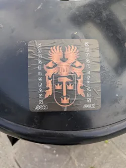 A rectangular sticker with rounded corners applied to a dark, possibly metal or plastic, surface. The sticker displays a stylized coat of arms or crest in a metallic copper-bronze color against a dark background. The central design features a shield containing a hammer and three crowns. Above the shield, there is a winged creature, likely an eagle, and intricate scrollwork flanks the main emblem. The words "KUNGENSBARN.COM" are printed vertically along both the left and right sides of the crest.