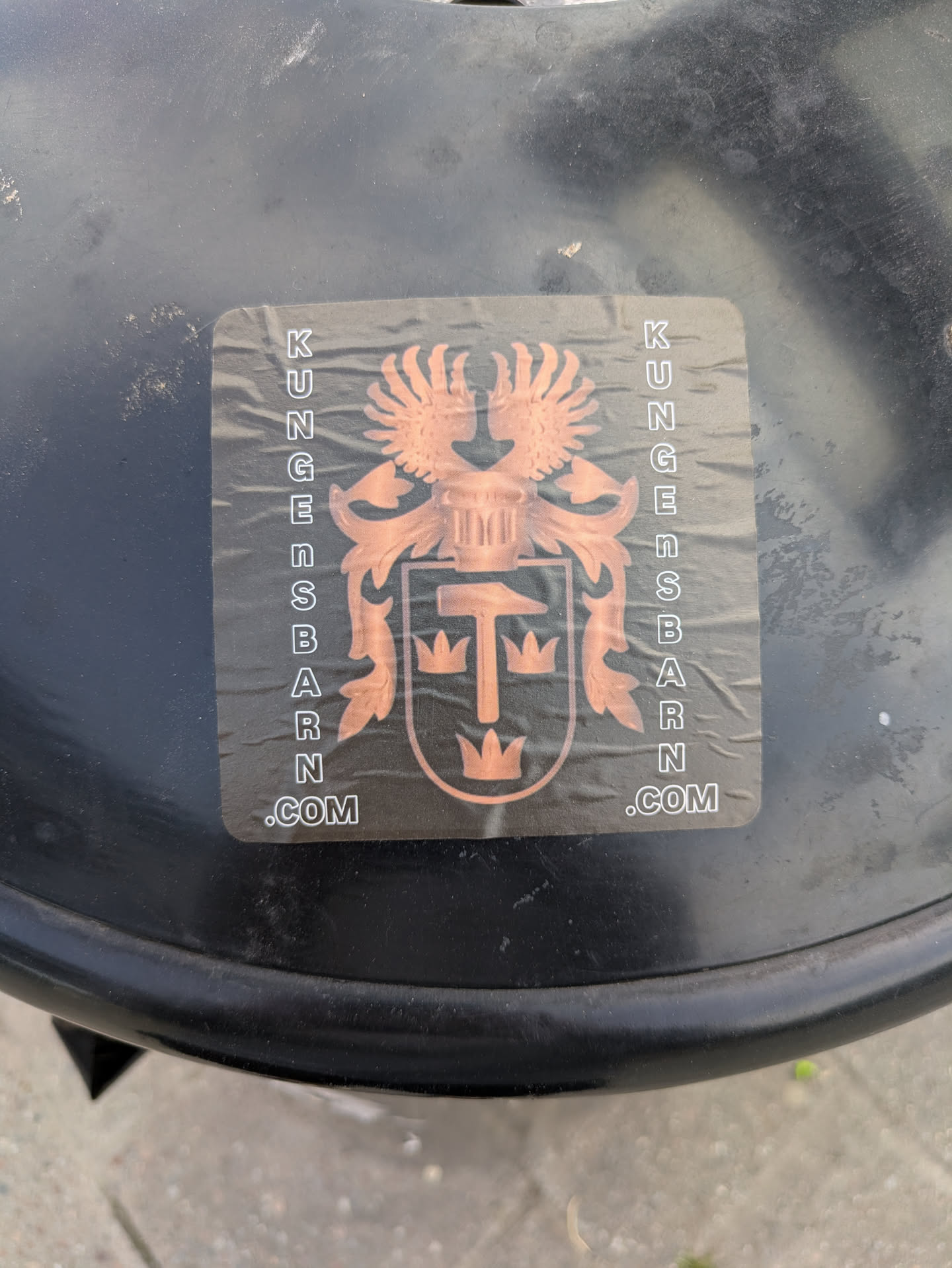 A rectangular sticker with rounded corners applied to a dark, possibly metal or plastic, surface. The sticker displays a stylized coat of arms or crest in a metallic copper-bronze color against a dark background. The central design features a shield containing a hammer and three crowns. Above the shield, there is a winged creature, likely an eagle, and intricate scrollwork flanks the main emblem. The words "KUNGENSBARN.COM" are printed vertically along both the left and right sides of the crest.
