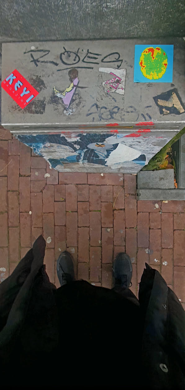 A collection of stickers, graffiti and torn posters are adhered to a metal surface. The leftmost sticker has red background and white text. Some of the stickers are partially damaged. Some graffiti is seen on top of the stickers.