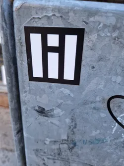 A square black and white sticker with a simple geometric design. The sticker is affixed to a weathered metal surface.
