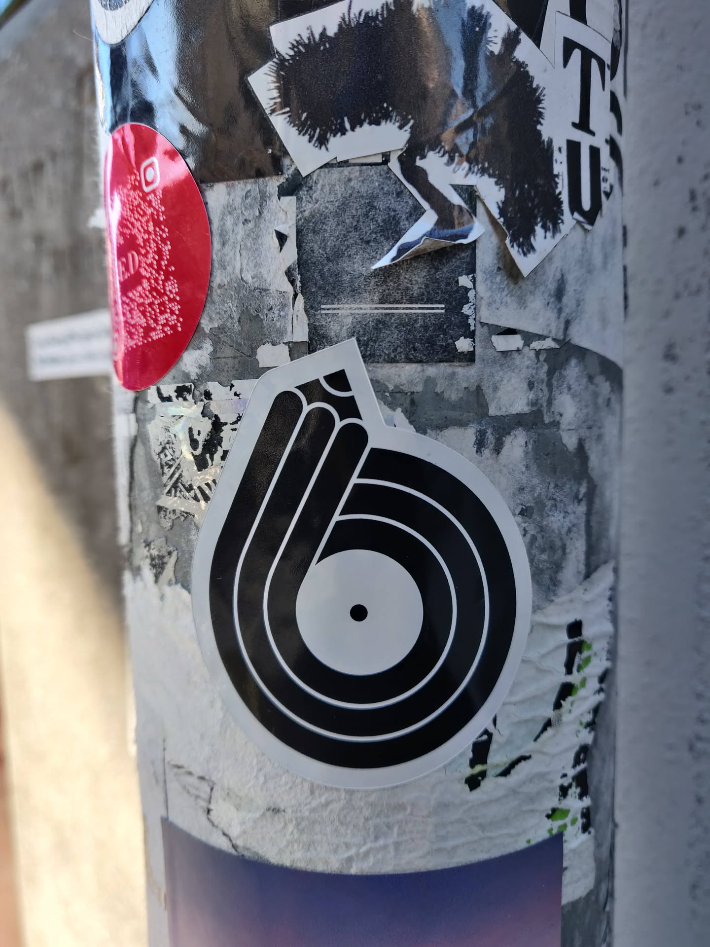 Black and white circular sticker with concentric circles reminiscent of a record or a stylized eye.  The central circle has a small dot in the middle.  The sticker is affixed to a pole covered in other stickers.