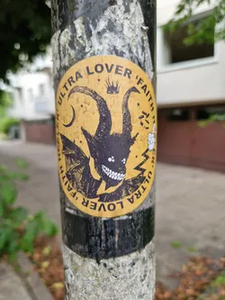 A round sticker featuring a demonic figure with horns and wings. The background is yellow, and the text 'Ultra Lover Faith' is written around the edge. There are other small details, like stars and a crown, in the background. The sticker is attached to a weathered pole.