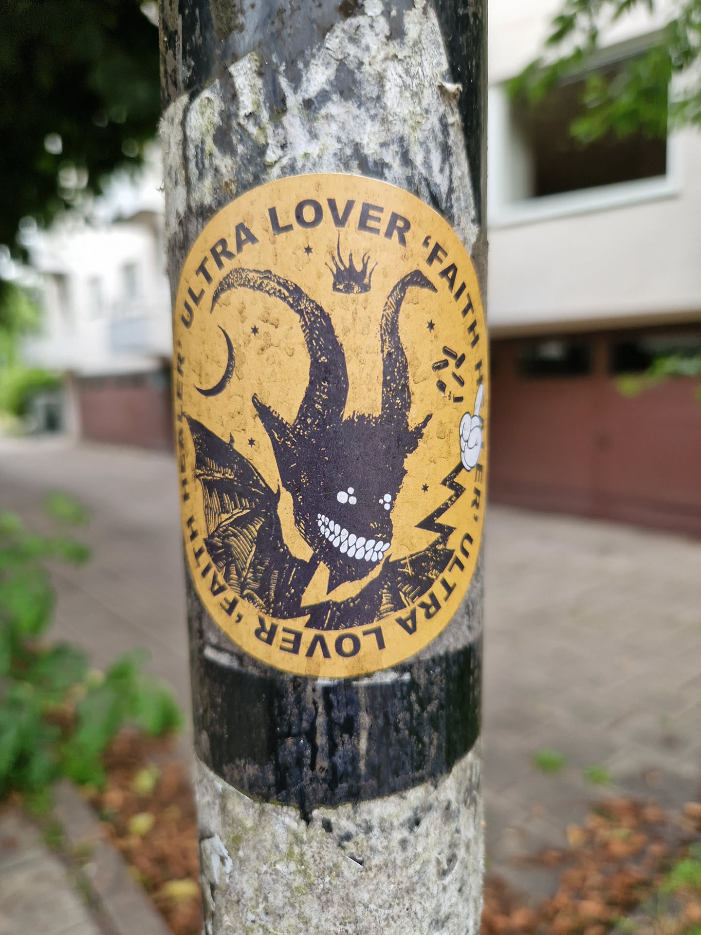 A round sticker featuring a demonic figure with horns and wings. The background is yellow, and the text 'Ultra Lover Faith' is written around the edge. There are other small details, like stars and a crown, in the background. The sticker is attached to a weathered pole.