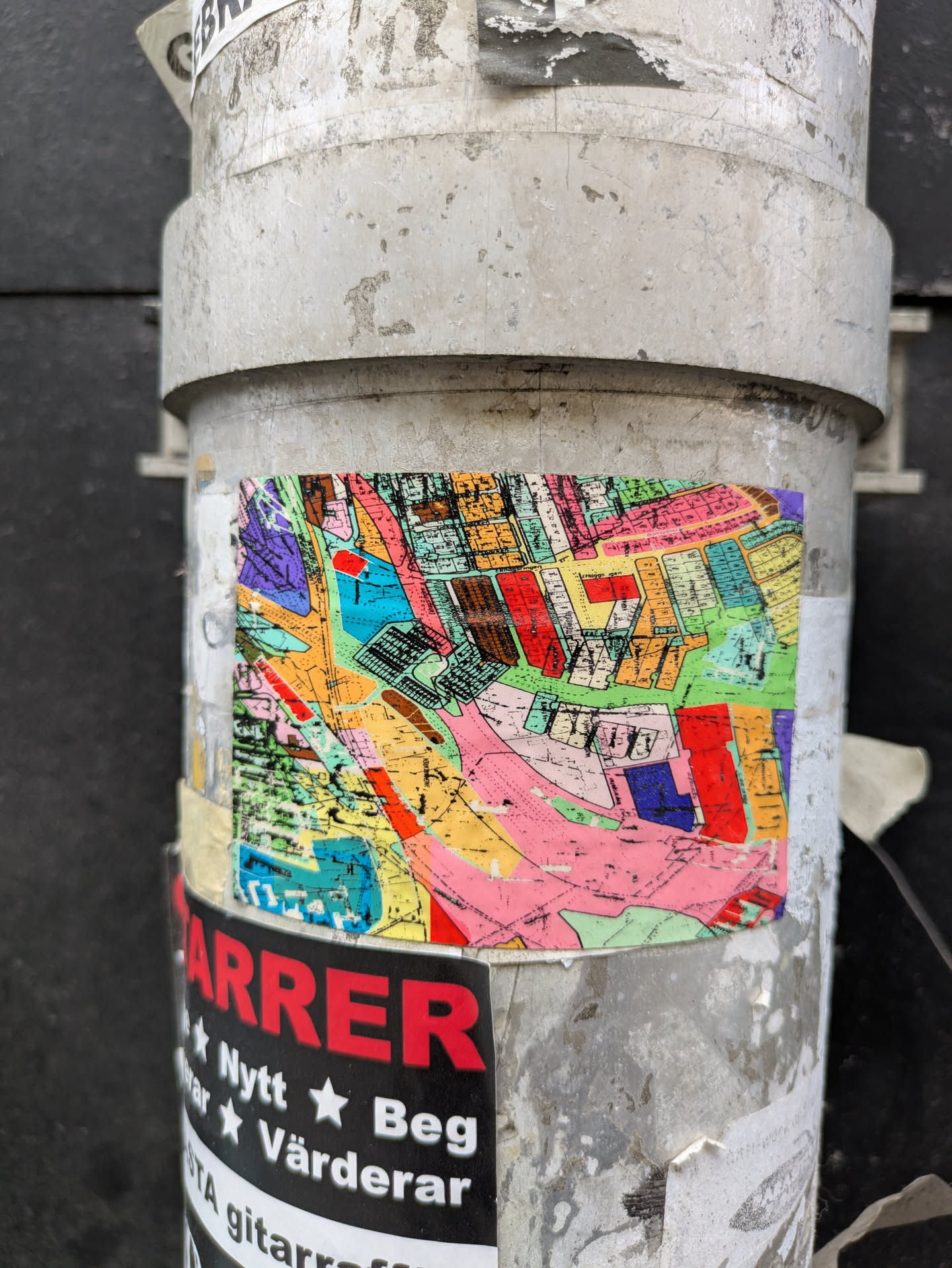 A sticker featuring a brightly colored map affixed to a cylindrical metal pole. The map appears to depict urban planning, with various shapes and colors representing different zones. The color scheme includes reds, blues, pinks, and yellows. The pole is weathered and has remnants of other stickers.