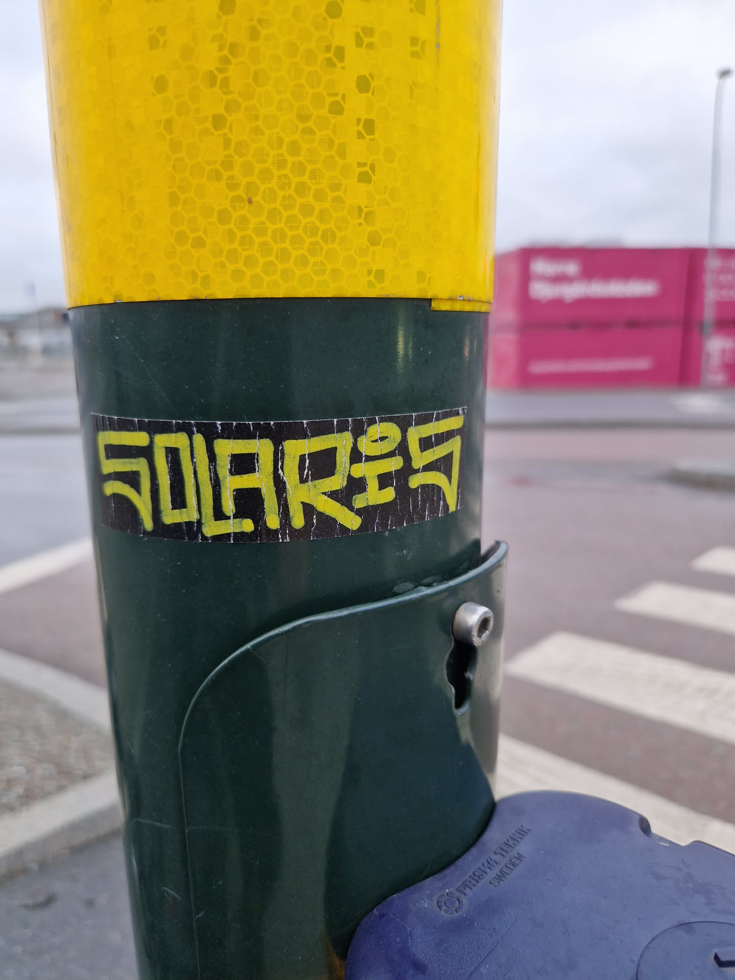 A black and yellow sticker with the word SOLARES written in a stylized font is affixed to a green pole. The pole appears to be part of a streetlight or similar urban fixture. The background is blurred but shows a pedestrian crossing and a pink building in the distance.